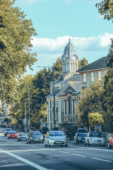 A street scene showing several parked and moving cars along a busy road in Bexley, with a historic church featuring a clock tower and a pointed spire in the background. The church has arched windows and decorative stonework, surrounded by tall leafy trees providing partial shade. In the foreground, a sidewalk lined with trees and lampposts runs parallel to the road, and a building with multiple windows is partially visible behind the trees. The image captures a clear, sunny day with a bright blue sky and scattered clouds, illustrating an urban environment that may be involved in house removals, with trucks or vehicles from Man with Van Bexleyheath potentially involved in the logistics of furniture transport or home relocation services.