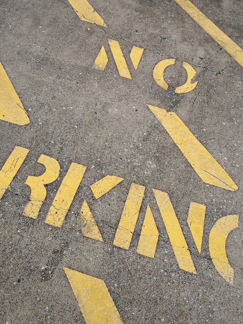 Close-up view of a grey asphalt pavement surface displaying faded yellow markings including the word 'NO' and partially visible directional arrows, indicating parking restrictions or traffic instructions in a residential or urban area. The markings are worn, showing signs of weathering and use. The image likely captures a designated zone relevant to house removals or moving logistics, such as a no-parking zone or loading area, supporting the context of planning a move or furniture transport. The scene is outdoors with no additional objects or vehicles visible, emphasizing surface details and the condition of the painted markings as they relate to urban parking regulations managed by Man with Van Bexleyheath for house removals services.