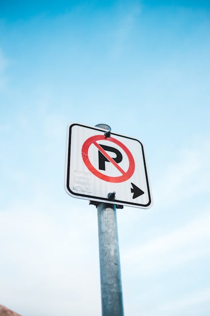 A vertical metal pole supporting a rectangular white traffic sign with a black border, indicating no parking with a red circle and diagonal line crossing out a black letter 'P', and an orienting black arrow pointing to the right. The sign is mounted against a bright blue sky with a few wispy clouds. The image captures a clear outdoor setting, typical of urban areas where house removals and furniture transport may occur, with the sign potentially indicating parking restrictions relevant to moving logistics. The sign’s placement on the pavement suggests a location near a property entrance or street, highlighting the importance of checking local parking rules during home relocation or moving processes, an aspect that Man with Van Bexleyheath helps clients manage through professional removals services.