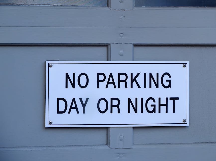 A rectangular white metal parking sign with bold black text reading 'NO PARKING DAY OR NIGHT' mounted on a light grey exterior wall of a building or garage. The sign is secured with four screws, one at each corner. The background surface has horizontal wooden paneling with visible nail heads and a slight texture, characteristic of a residential or commercial property. The image is well-lit with natural light, highlighting the sign's clarity and the surface details around it. This visual relates to house removals and relocation logistics, where clear signage can help avoid parking fines during furniture transport and home relocation processes, and is relevant for services like those offered by Man with Van Bexleyheath, supporting efficient packing, loading, and moving activities.