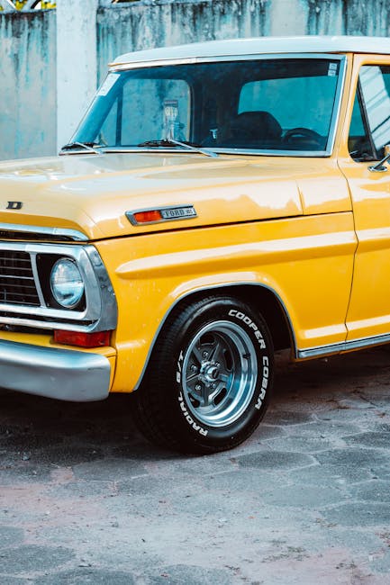 Close-up of a vintage yellow Ford D-series truck parked outdoors, with a focus on the front left side. The vehicle features a rectangular headlamp, a chrome bumper, and a black grille. The bodywork shows some slight wear, indicative of age, with visible panel lines and a side badge. The front wheel, fitted with a black Cooper Cobra tire and a silver rim, is turned slightly to the right. The truck is positioned on a paved surface, with a weathered, greenish wall or fence in the background. This image exemplifies classic vehicle preservation, often associated with home relocation or furniture transport services, as part of the detailed process of packing and moving for house removals, facilitated by professionals like Man with Van Bexleyheath.