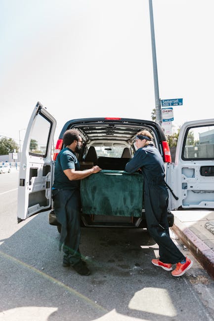 Two men wearing dark uniforms and protective glasses are involved in a house removal process, carefully loading a large, dark green upholstered armchair into the open back of a white van parked on a street. One man is holding the armchair steady while the other guides it into the vehicle, which is part of a furniture transport or packing and moving operation. The van's interior reveals a spacious cargo area, and the surrounding environment includes a residential street with a sidewalk and a street lamp visible in the background, along with clear daylight. This scene demonstrates the loading process typical of home relocation services offered by Man with Van Bexleyheath, involving the handling of bulky items, packing materials, and transportation equipment in preparation for a house move.