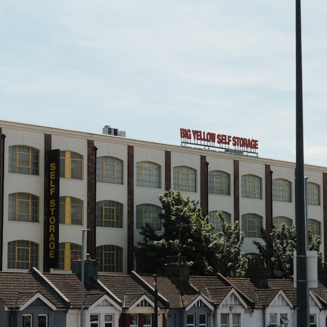 A multi-storey building with a white facade and multiple large, yellow-framed windows, marked with a black vertical sign reading 'Self Storage' on the side. The building features brick accents between the windows and is situated behind a row of small residential houses with tiled roofs. In front of the building, several trees partially obscure the view, and a street lamp is visible on the right side of the image. The scene is captured during daylight with clear weather, and a sign on the roof reads 'Big Yellow Self Storage.' The setting suggests a commercial property used for storage services, which may relate to house removals and packing logistics managed by [COMPANY_NAME], helping clients with home relocation and furniture transport.