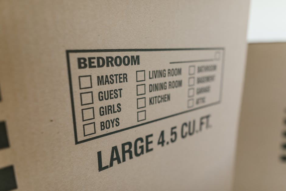 Close-up of a brown cardboard moving box with printed section labels for different rooms such as bedroom, living room, dining room, and kitchen, each with small checkboxes. The bedroom section includes options for master, guest, girls, and boys, with some boxes checked or unchecked. The box is marked with the size description 