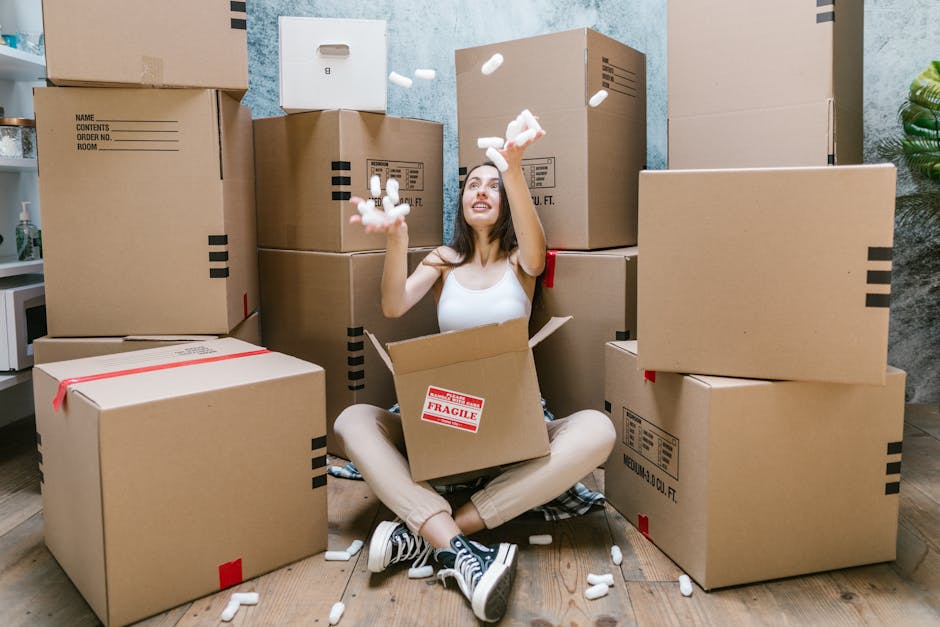 A woman sitting on the floor inside a room surrounded by numerous large brown cardboard moving boxes, some sealed with red tape and others labeled with contents and handling instructions. She is wearing casual clothing, including a white tank top and striped sneakers, and is happily throwing packing foam peanuts into the air from an open box that has a 'Fragile' sticker on it. The room has a wooden floor and a blue textured wall, with visible domestic items such as a white microwave on a small shelf and a potted green plant visible on the right side. This scene depicts the packing and moving process involved in home relocation, with the woman actively engaged in packing or unpacking, and the boxes arranged in a somewhat organized manner, suggesting a move managed by a professional removals service like Man with Van Bexleyheath.