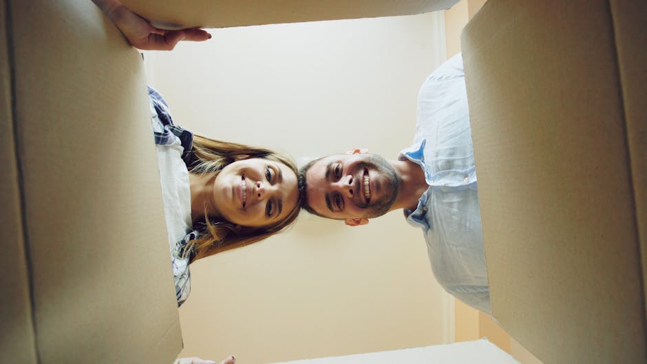 A man and a woman are seen from a low angle inside a room, lying on their backs beneath a large cardboard box with open flaps. The woman has long brown hair and is smiling, wearing a casual plaid shirt, while the man has short hair, a beard, and is also smiling, dressed in a light-colored casual shirt. They are positioned on what appears to be a carpeted floor, possibly during a house relocation process. The cardboard box, likely filled with household items or fragile belongings, is partially covering their bodies. The setting is well-lit with natural or bright indoor lighting, and the background shows plain cream-colored walls. The scene suggests a moving or packing activity related to home removals services, with the individuals preparing for or in the midst of a furniture transport or packing and moving process, as provided by Man with Van Bexleyheath.