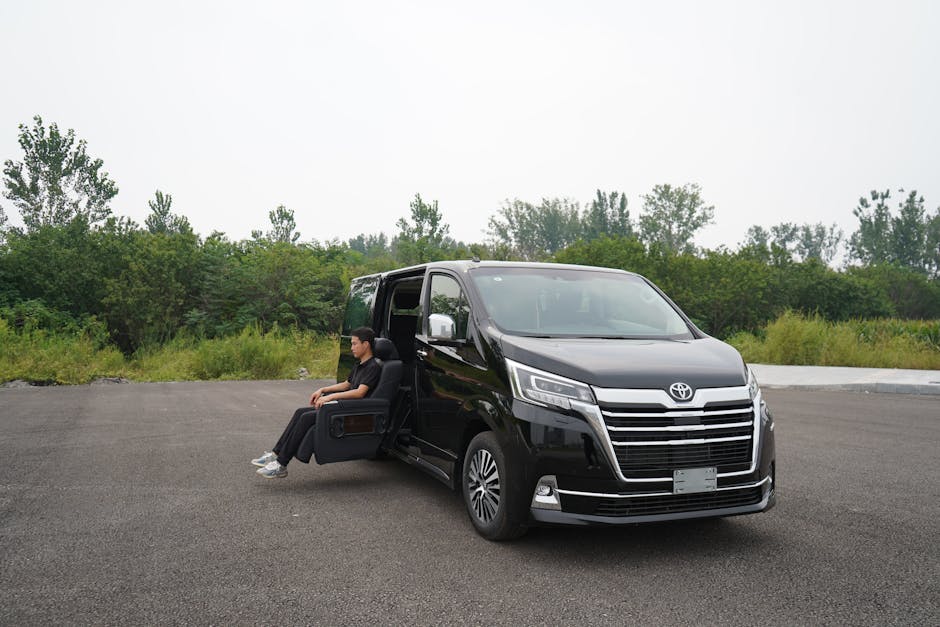 A person seated comfortably in a black armchair is partially outside a large black Toyota van, which is parked on an empty paved area surrounded by green trees and shrubs. The armchair is positioned with the backrest towards the open sliding side door of the van, suggesting an unusual loading or pre-move staging situation. The van is clean and modern, featuring a prominent front grille and silver accents, and appears to be used for home relocation or furniture transport services as part of a moving process. The environment is outdoors during daylight hours under overcast sky, with no other vehicles or objects in the immediate vicinity. This scene illustrates the logistics of loading or preparing furniture and belongings for a house delivery or move, highlighting the equipment and space used in professional removals, such as those by Man with Van Bexleyheath.
