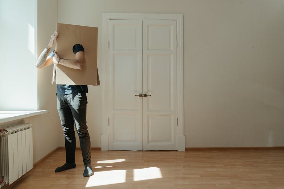 A person holding a large cardboard box against their shoulder inside a room with wooden flooring and plain walls. The individual, dressed in dark pants and a dark top, is standing near a window with natural light illuminating the space. The room has a white double door with a metallic latch, and a white radiator is visible below the window. The scene depicts the process of packing and preparing for home relocation, with the person likely involved in moving or furniture transport activities. This image reflects the packing phase of house removals, supported by the presence of the cardboard box. Man with Van Bexleyheath, a professional removals service, would assist with such packing and furniture transport tasks during small flat moves in Bexley Village, as described on their website.
