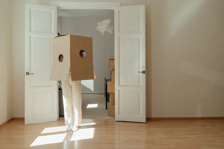 A person dressed in beige trousers and a long-sleeved shirt, carrying a large cardboard box with circular cutouts and a protective white blanket wrapped around their legs, is walking through a set of open white double doors into a room. Inside the room, there are stacked moving boxes of various sizes, some made of cardboard with packing tape. The boxes are positioned on a black table or surface, and a crumpled piece of packing paper is hanging from the ceiling. The room has light-colored wooden flooring and neutral beige walls. Sunlight streams in through a window, casting bright patches of light on the floor. The setting depicts a home during the process of packing and furniture transport as part of house moves, with the individual actively involved in a furniture transport or packing and moving activity for a professional removals service like Man with Van Bexleyheath.