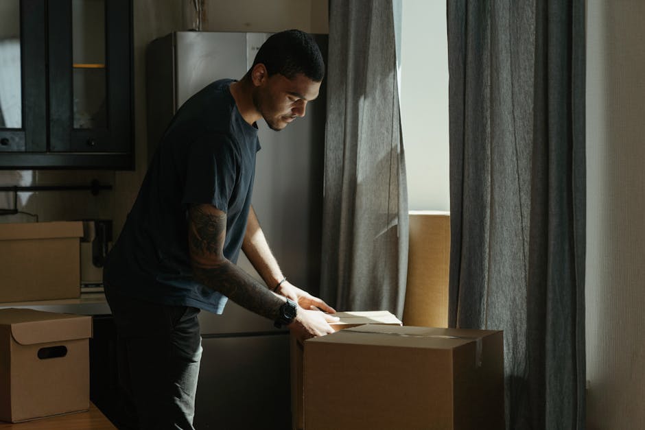 A person holding a large cardboard box against their shoulder inside a room with wooden flooring and plain walls. The individual, dressed in dark pants and a dark top, is standing near a window with natural light illuminating the space. The room has a white double door with a metallic latch, and a white radiator is visible below the window. The scene depicts the process of packing and preparing for home relocation, with the person likely involved in moving or furniture transport activities. This image reflects the packing phase of house removals, supported by the presence of the cardboard box. Man with Van Bexleyheath, a professional removals service, would assist with such packing and furniture transport tasks during small flat moves in Bexley Village, as described on their website.