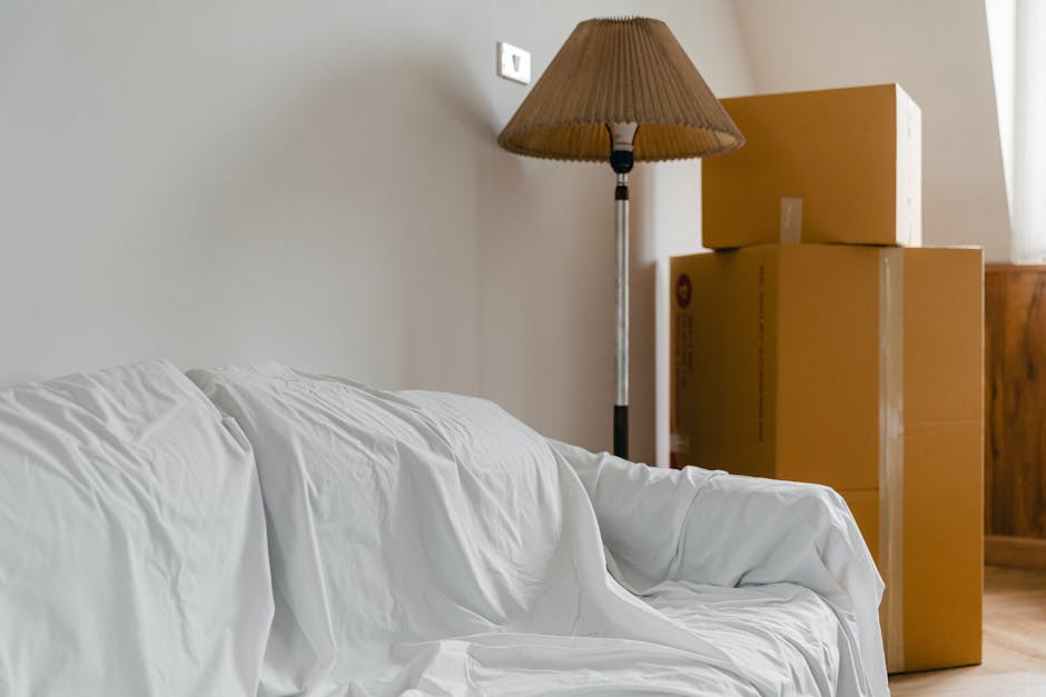 Inside a residential room with a white fabric-covered sofa positioned on the left side of the image, showing slight creases on the material. Behind the sofa, a wooden sideboard is partly visible with three large cardboard moving boxes stacked on top—two medium-sized boxes and one larger box, all sealed with clear packing tape. To the right of the boxes, a tall floor-standing lamp with a brown pleated lampshade and a metallic stand is present, casting soft light on the scene. The background includes a beige or light-colored wall with a white electrical socket near the top left of the image. Natural light illuminates the room from an unseen source, highlighting the packing and furniture items, indicative of the ongoing process of home relocation or furniture transport carried out by [COMPANY_NAME], supporting the packing and moving phase of house removals.