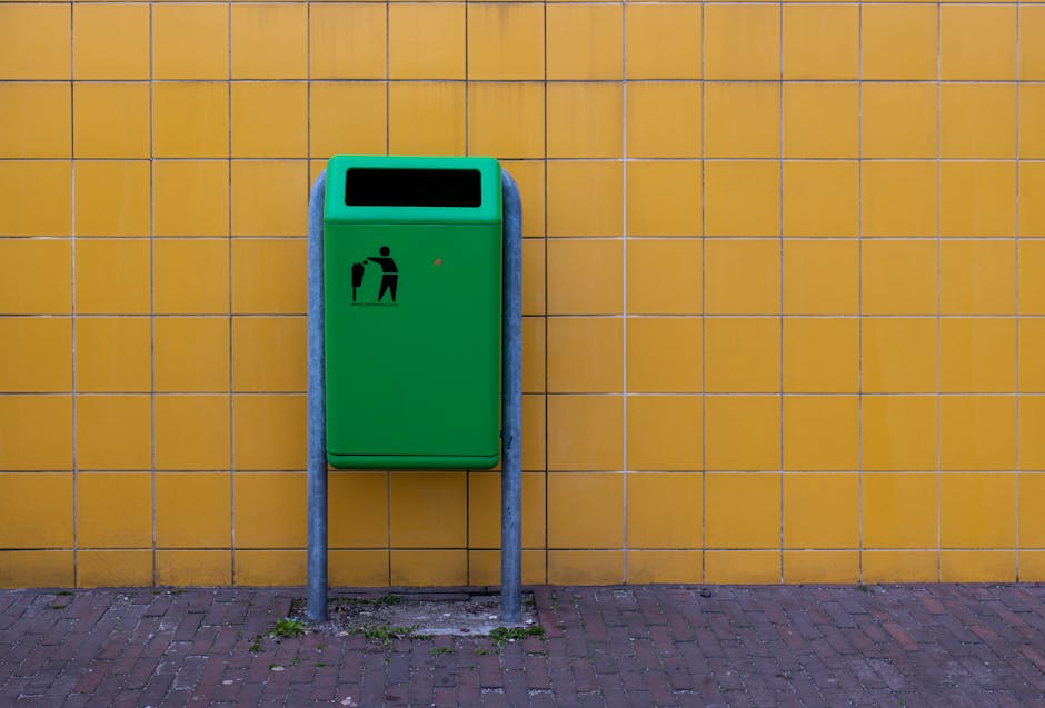 A row of four waste disposal bins are positioned along a cobblestone pavement in front of an orange building with barred windows and an arched doorway. The first bin on the left is dark green with a g